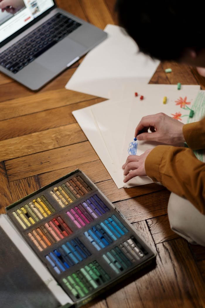 contact-img Adult engaged in online art class, using oil pastels on wooden floor.